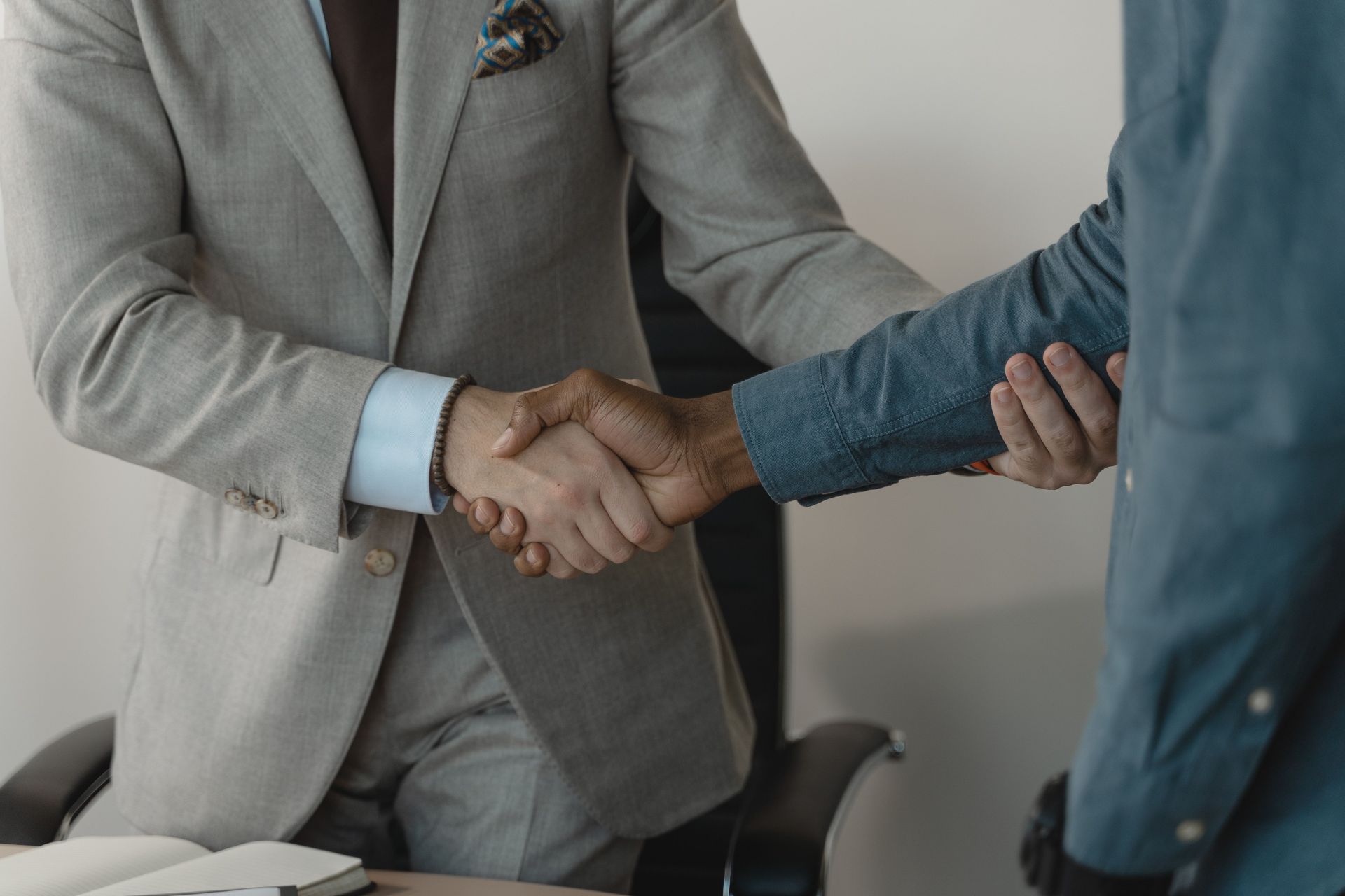 Two people shake hands across a desk, one in a gray suit and one in a denim shirt.