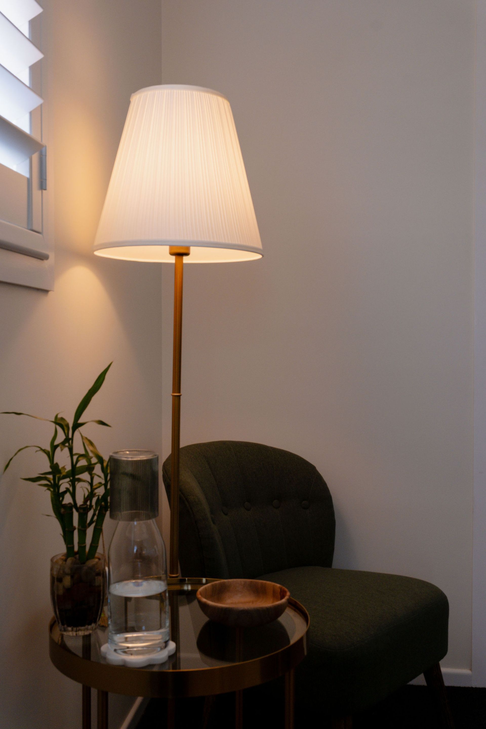 Cozy corner with a lit floor lamp, dark chair, side table, and potted plant by a window