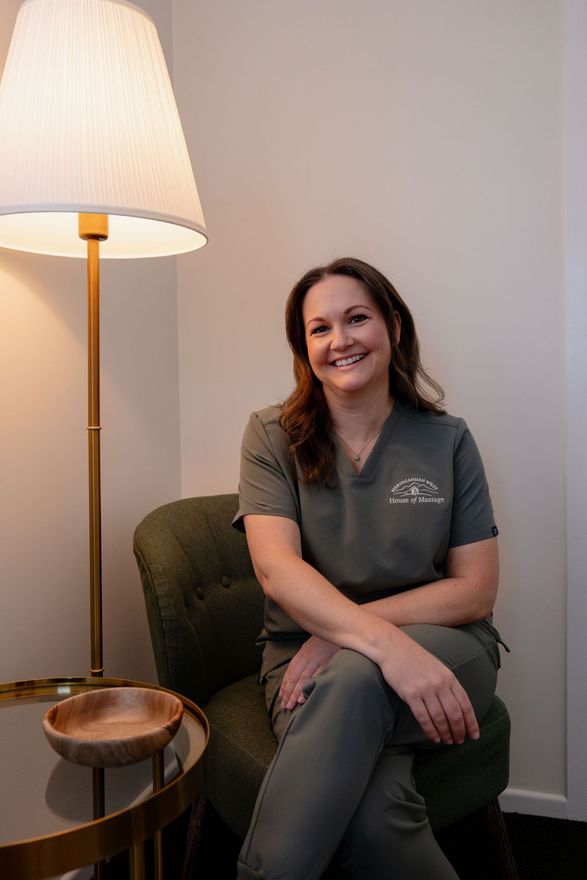 Woman in gray scrubs seated in a chair beside a lit floor lamp in a quiet room.