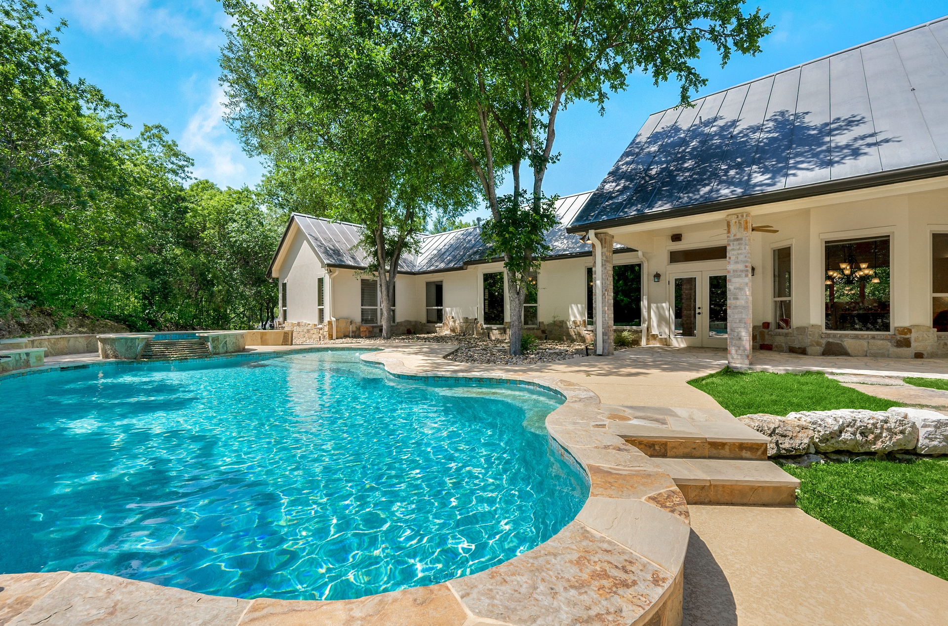 Luxurious home with a pool: blue water, stone patio, green lawn, trees, white house with dark roof.