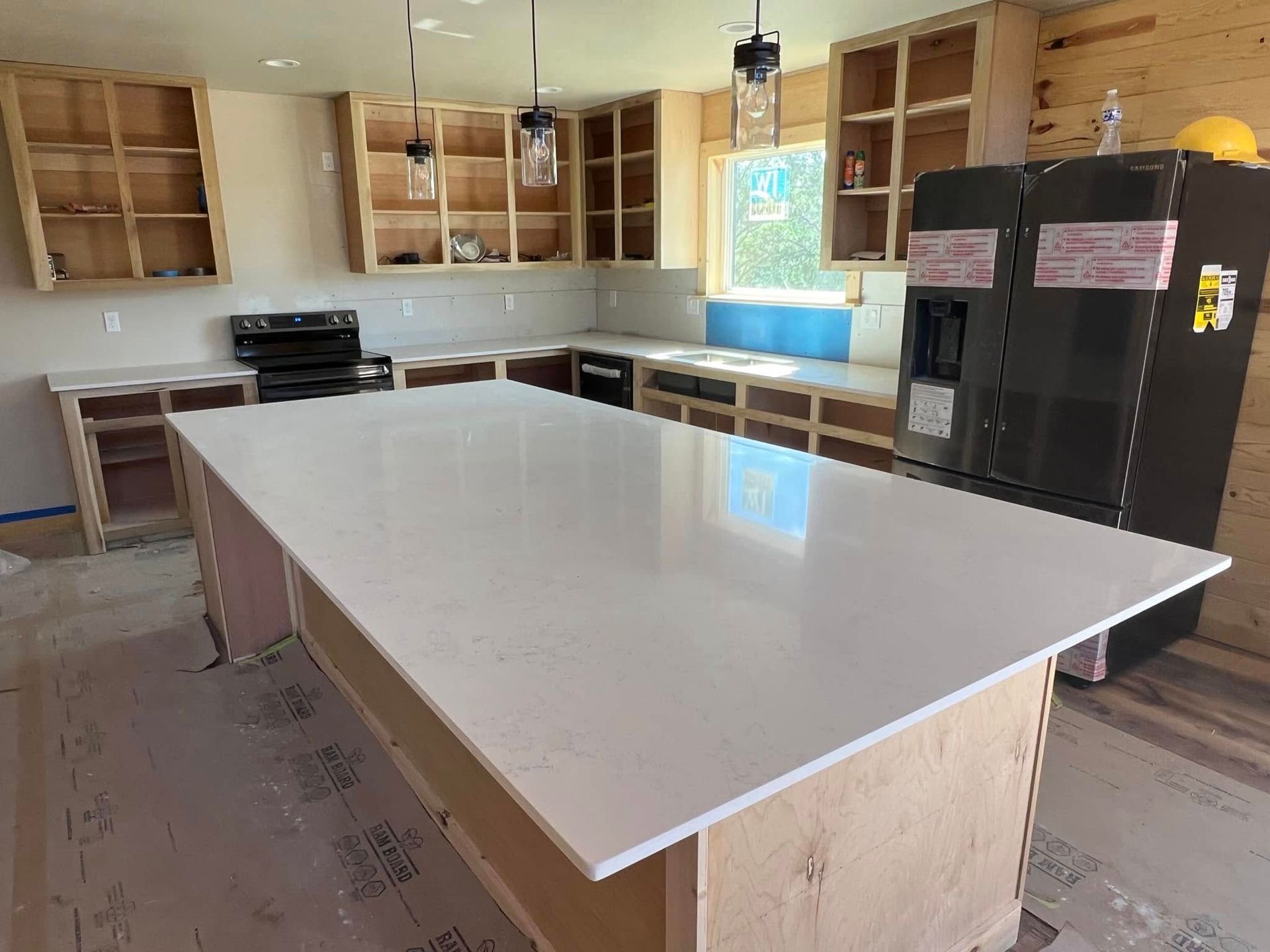 Unfinished kitchen with large white island, open cabinets, black refrigerator, and windows above the sink