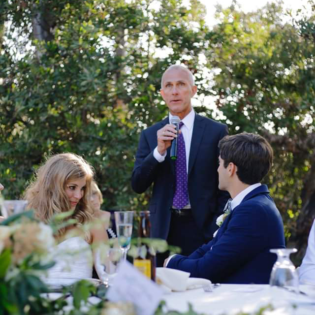 A man in a suit and tie is speaking into a microphone at a wedding reception