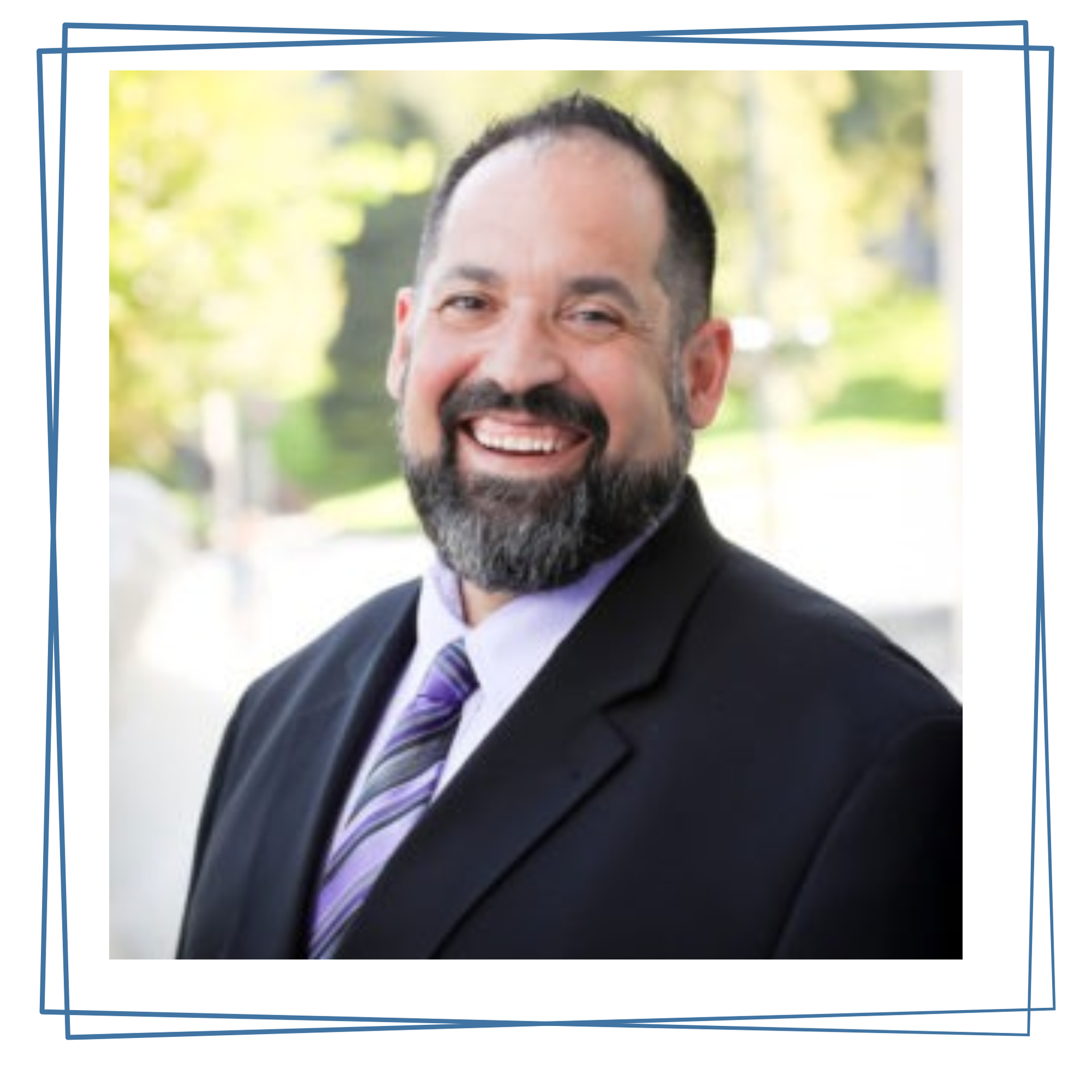 A person with a beard, wearing a suit and purple-striped tie, smiling outdoors in a headshot portrait.
