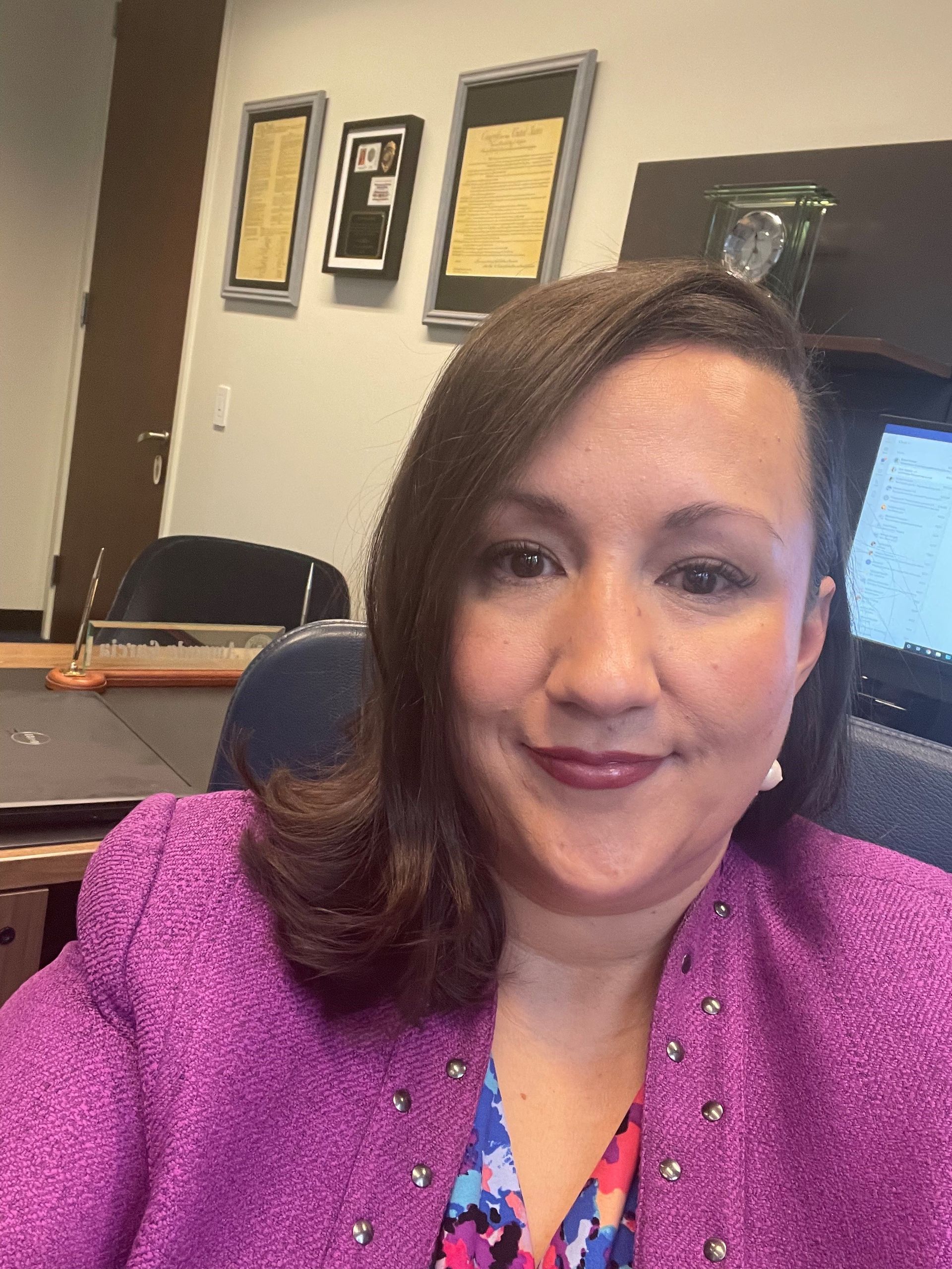 Woman in a pink blazer smiles at the camera in an office with framed documents.