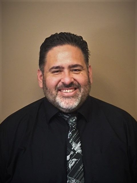 A man with a beard and glasses wearing a suit jacket and collared shirt smiles in front of a wood-paneled wall.