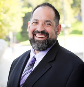 Smiling man in a dark suit and purple tie outdoors, with a blurred green background