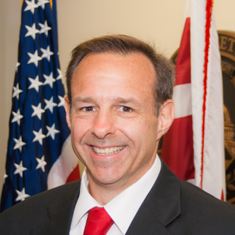 Man in suit with red tie smiles in front of US and Alabama flags.