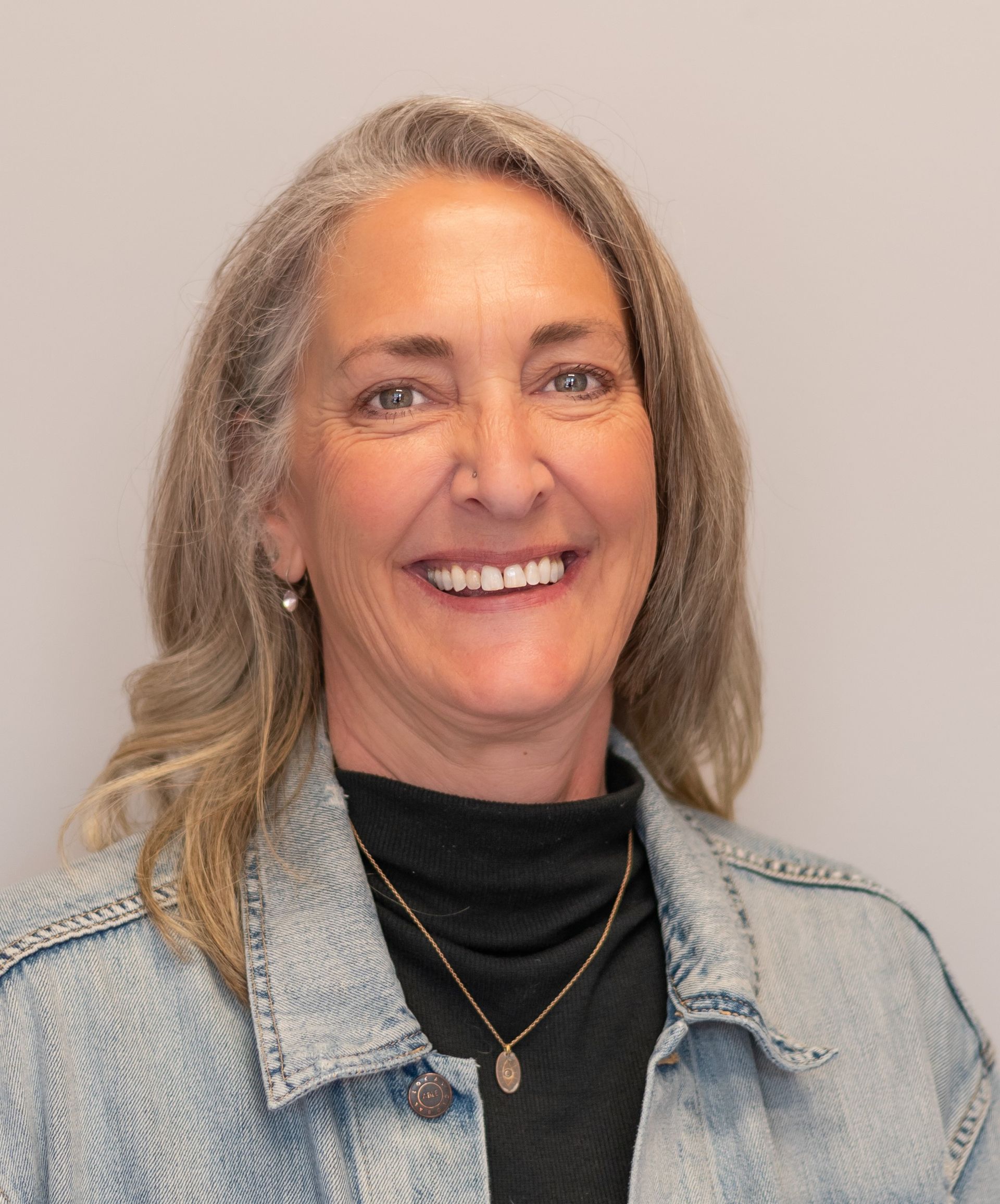 Woman with blonde hair, smiling, wearing a black turtleneck and denim jacket, gold necklace.