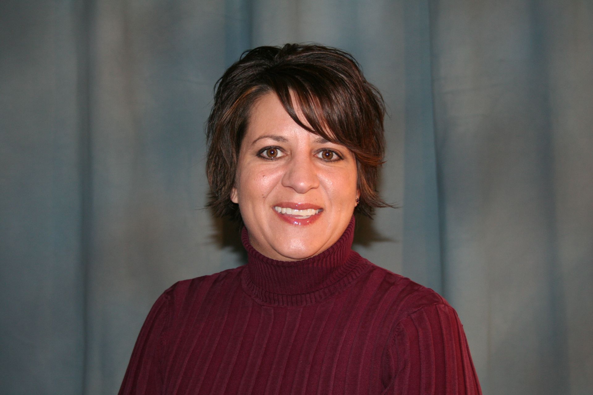 Woman with short brown hair smiles, wearing a burgundy turtleneck. Blue curtain backdrop.
