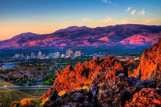 City skyline at sunset, mountains in background, orange and pink hues, rocky foreground.