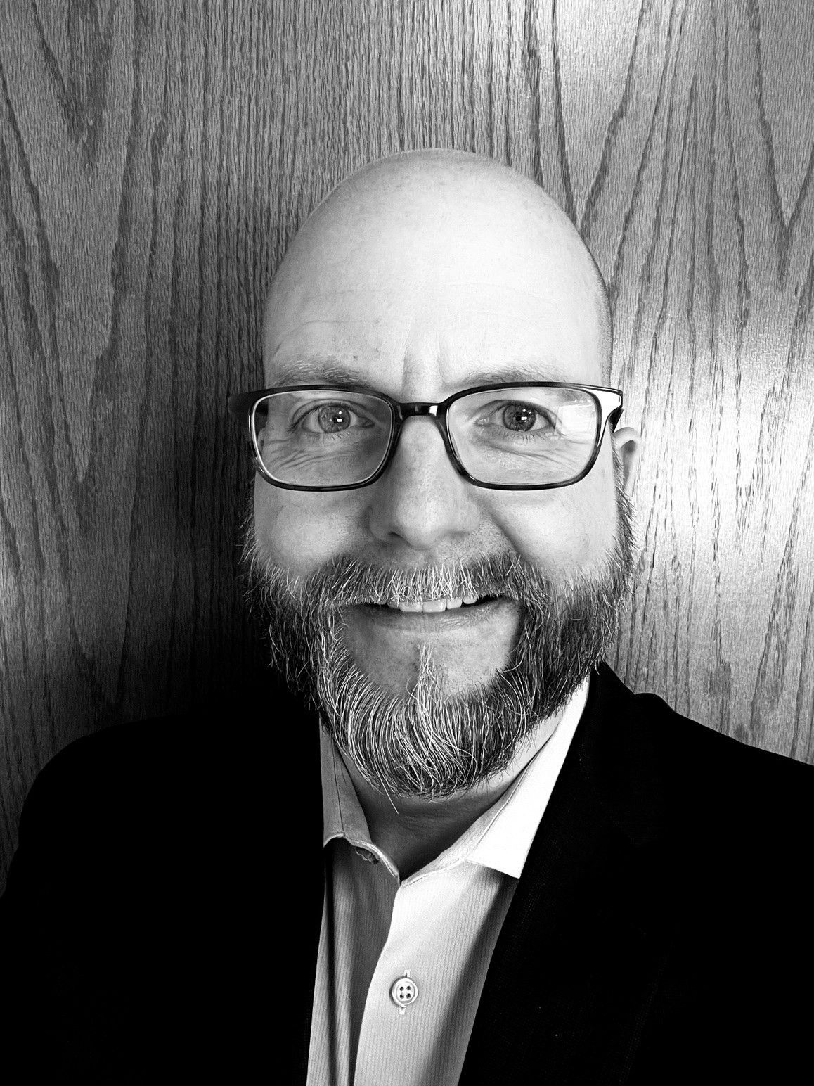 A man with a beard and glasses wearing a suit jacket and collared shirt smiles in front of a wood-paneled wall.
