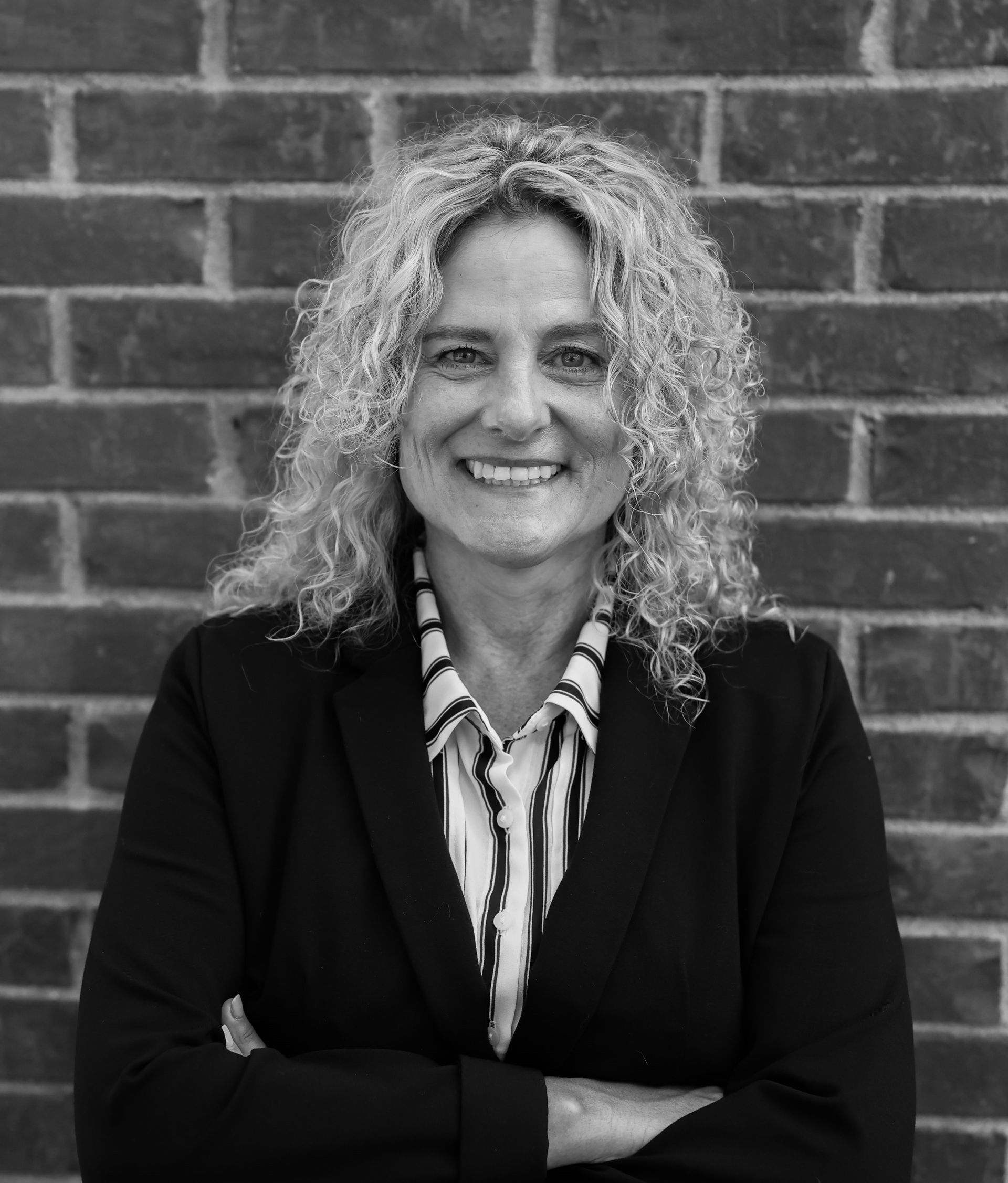 Woman with curly hair, arms crossed, smiles in front of a brick wall. She wears a blazer and striped shirt.