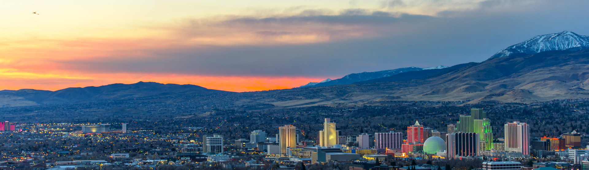Reno, Nevada skyline at sunset with mountain backdrop. Buildings are brightly lit, sky is orange and blue.