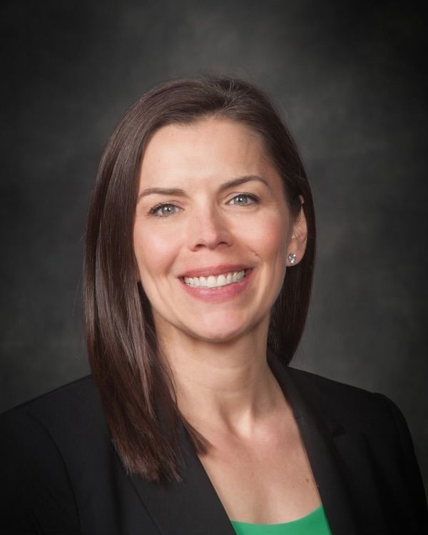 Woman in a black blazer smiles at the camera, dark hair, neutral background.