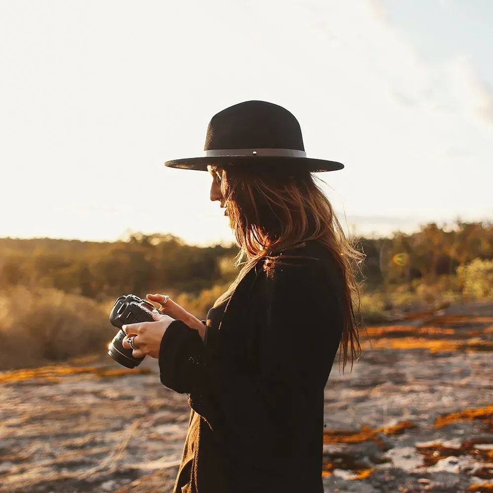 Woman in black hat and coat holds a camera outdoors, looking at the screen, with a background of trees.