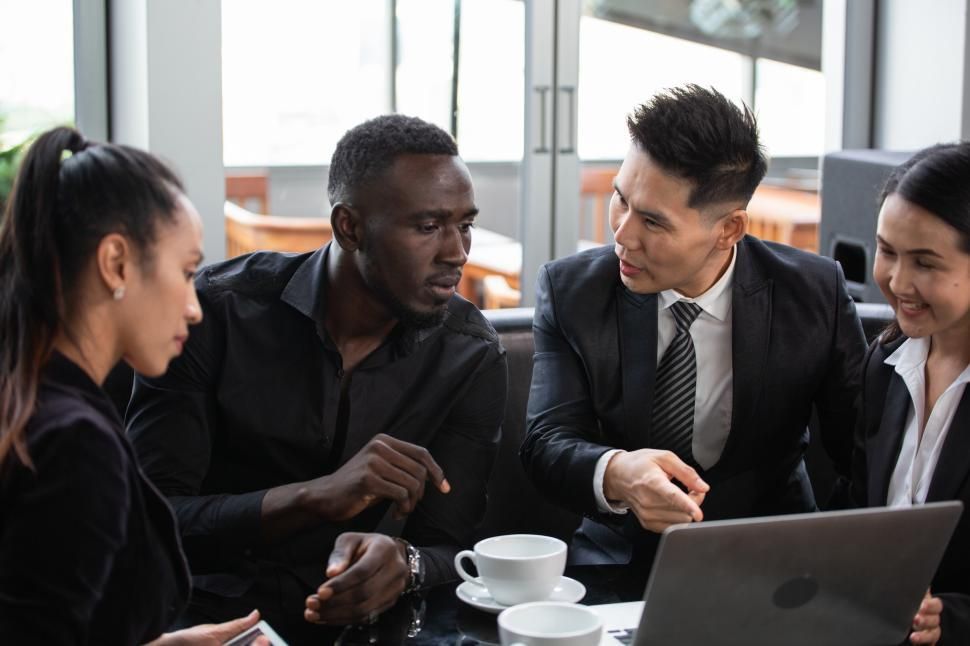 Four business professionals in suits reviewing a laptop together, focused discussion, light-filled interior.