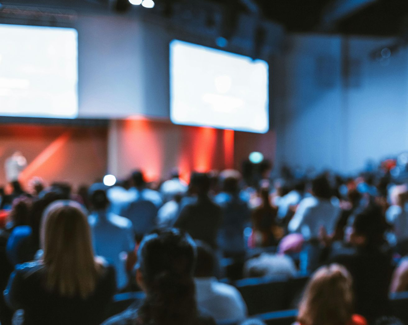 Audience seated in a dim conference hall facing bright presentation screens