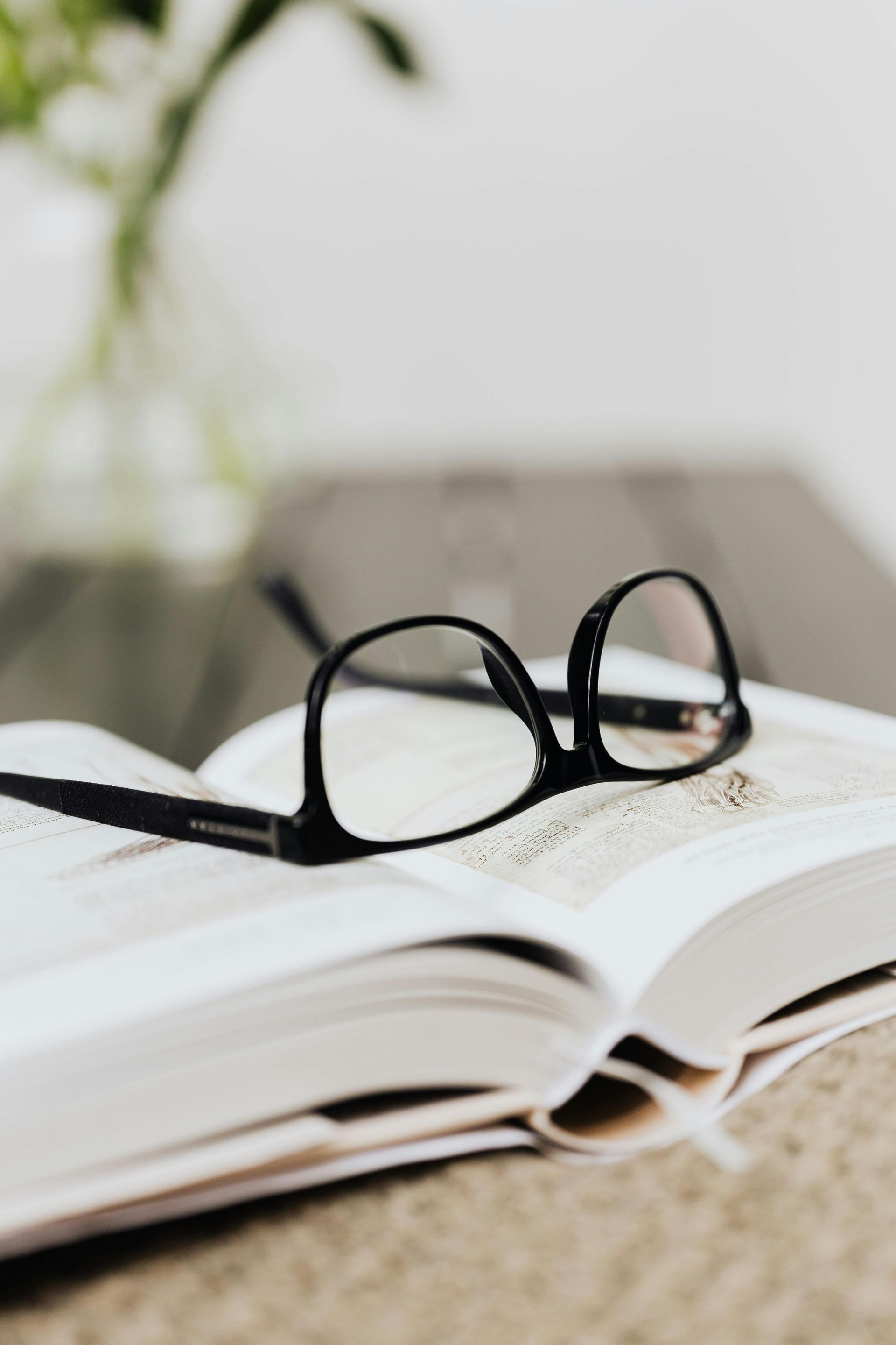 Black eyeglasses on an open book, blurry background with a plant.