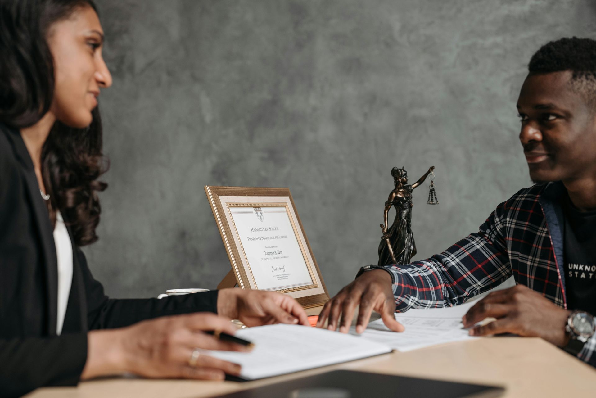 A person in a blazer consults with another person over documents at a desk, statue of Lady Justice.