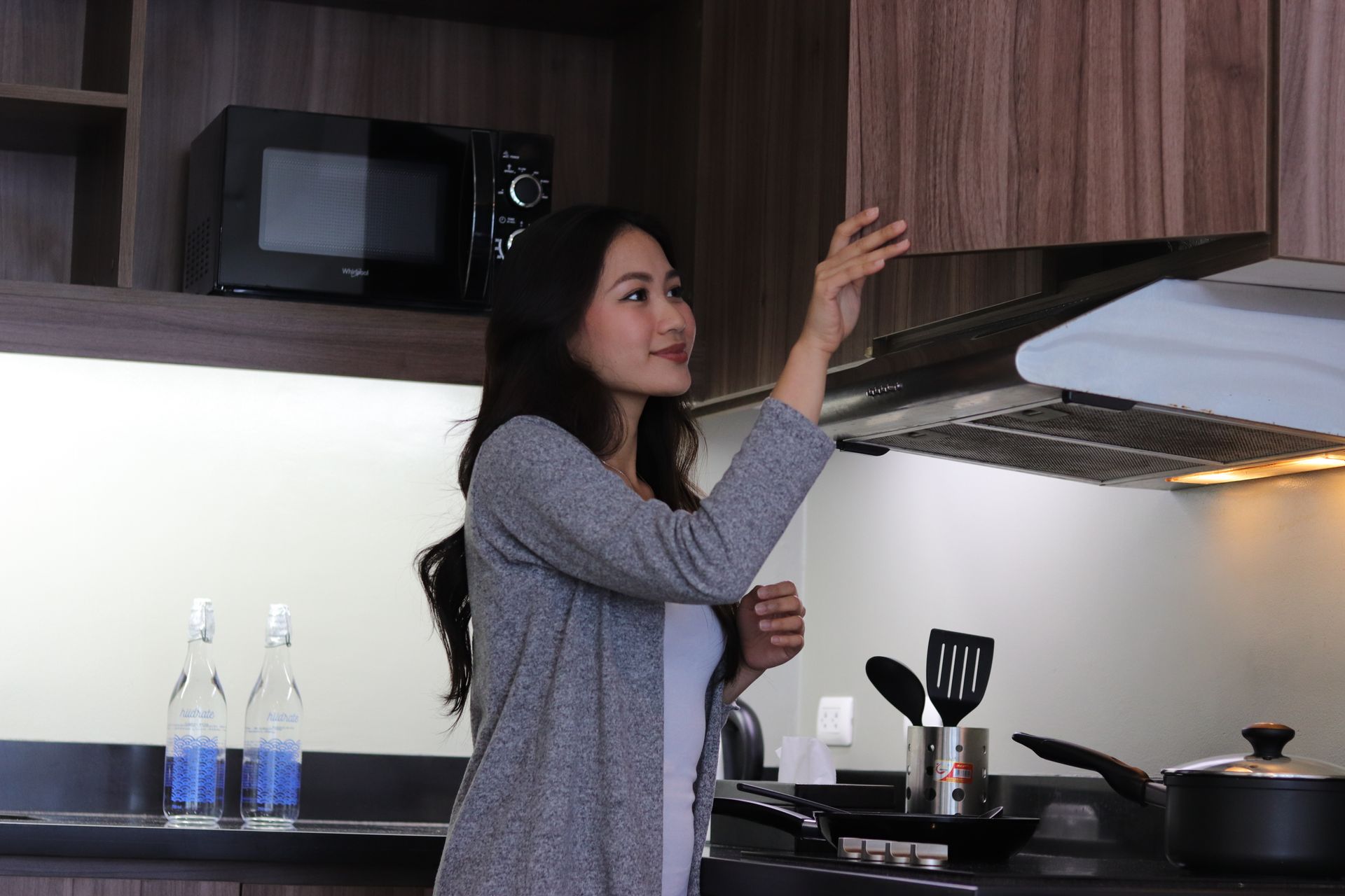 Woman reaching toward a kitchen range hood in a modern and spacious kitchen with a microwave and stove.