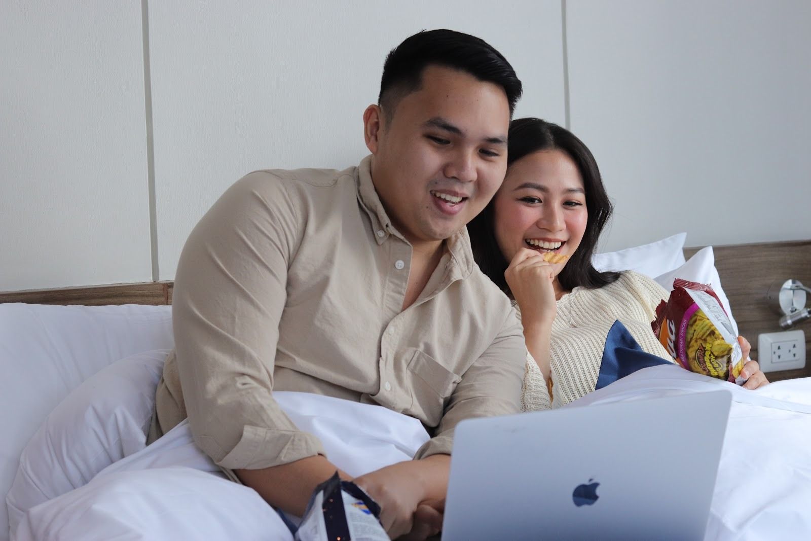 A man and a woman are sitting on a bed looking at a laptop.