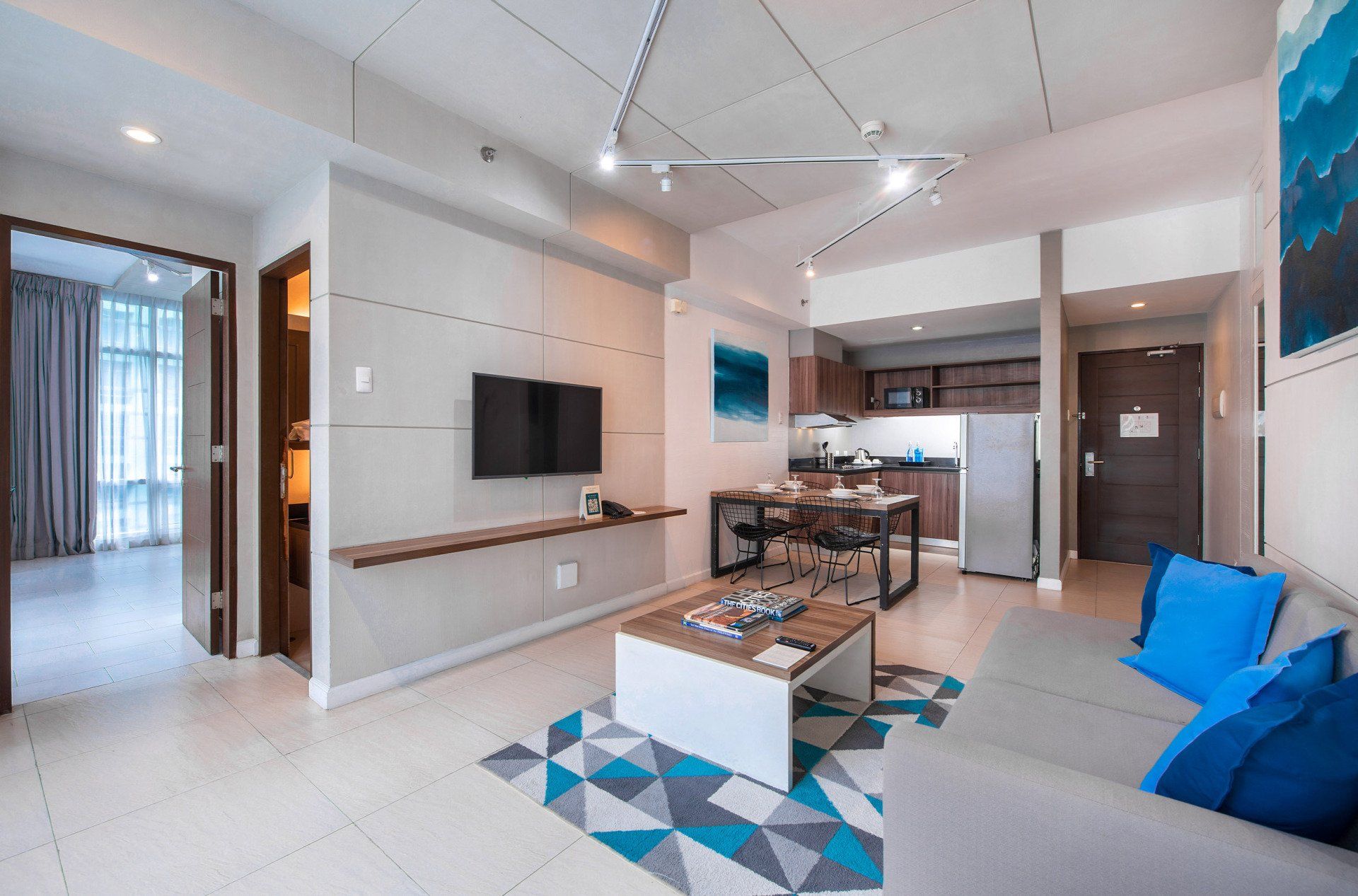Modern apartment interior with living area, kitchen, and dining table. Features gray couch, TV, and blue accents.