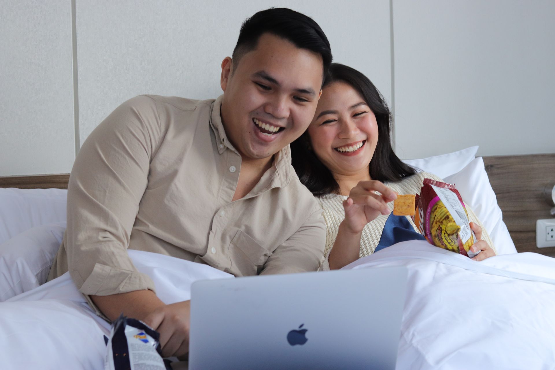 Couple laughing while watching a laptop and sharing snacks in bed at a serviced apartment at The Sphere Makati