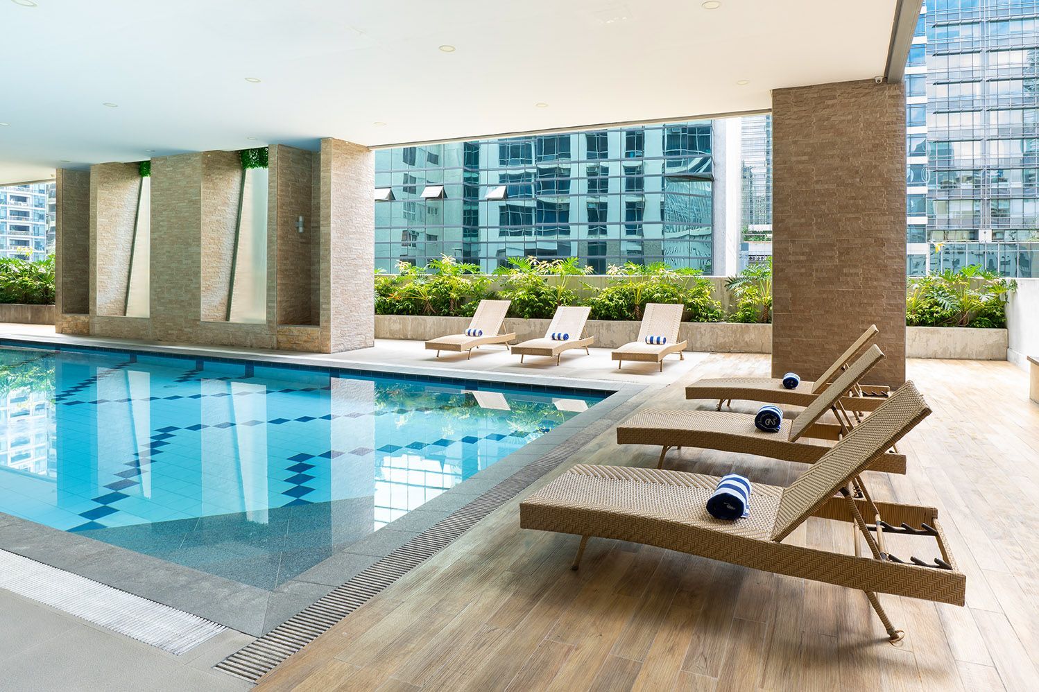 Indoor swimming pool with lounge chairs on a wooden deck and a backdrop of urban buildings.