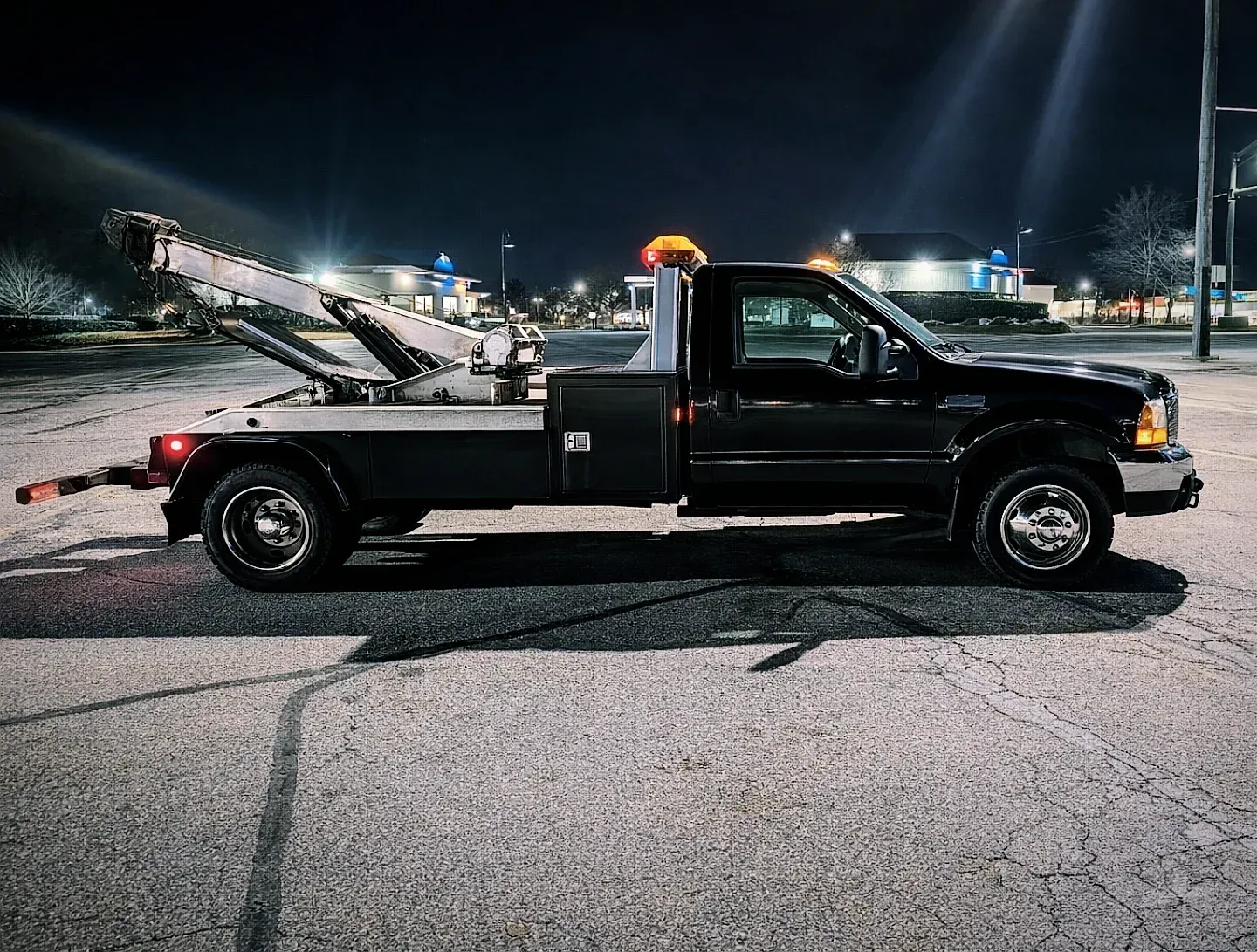 Black tow truck parked at night in a lit parking lot, side view