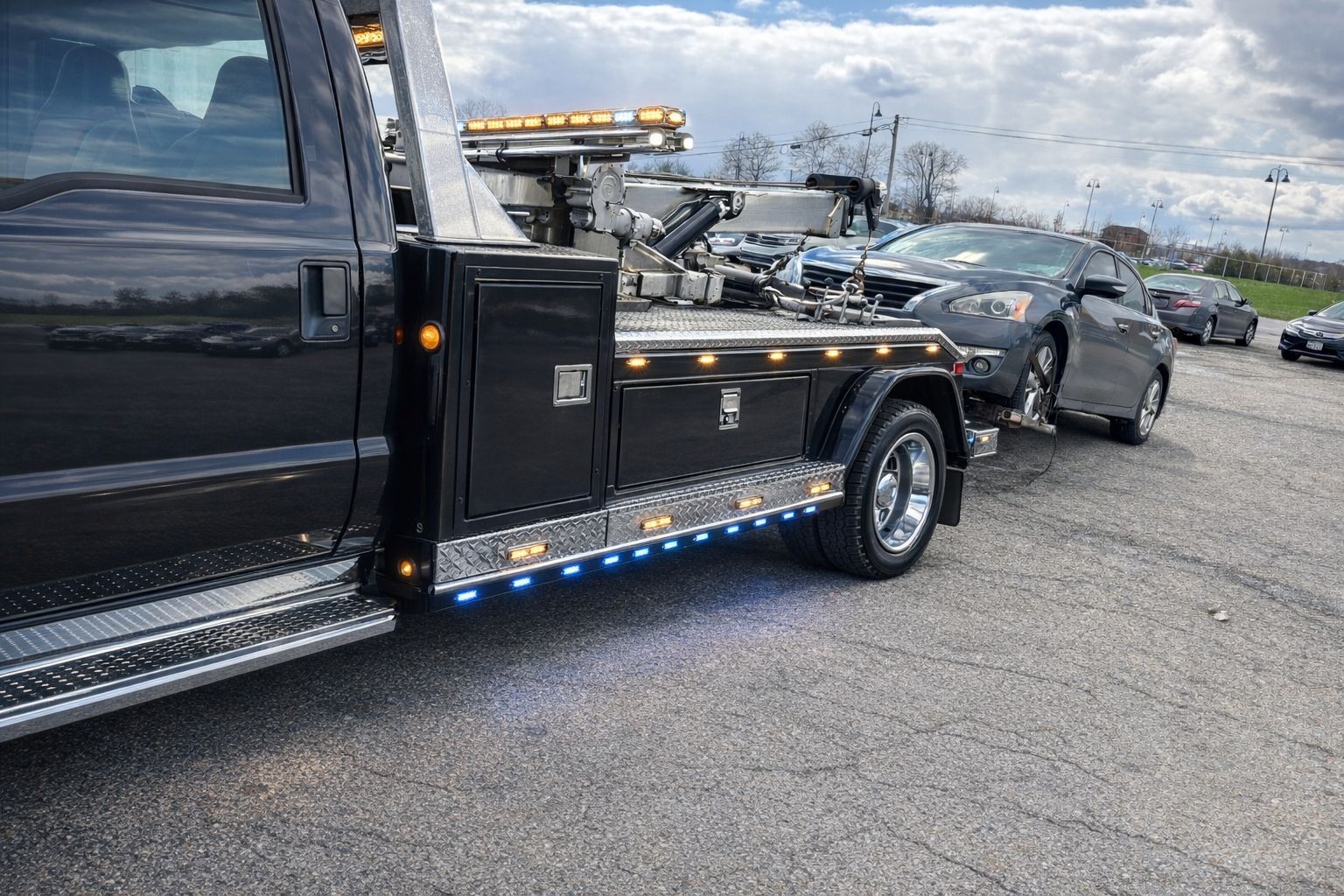 Black tow truck hauling a damaged silver sedan on a roadside under cloudy skies
