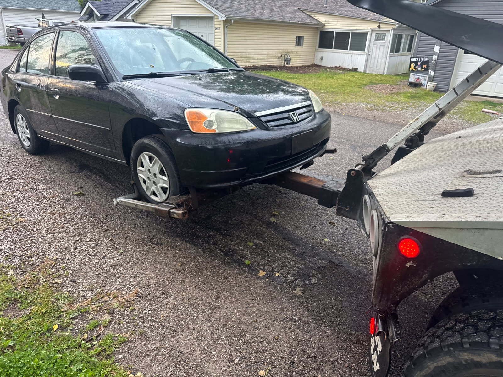Black sedan being towed by a flatbed truck on a wet driveway.