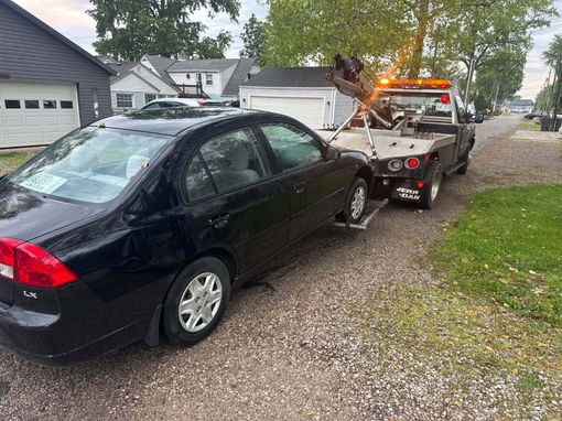 Black car being towed on a gravel roadside in front of houses, with tow truck lights on.