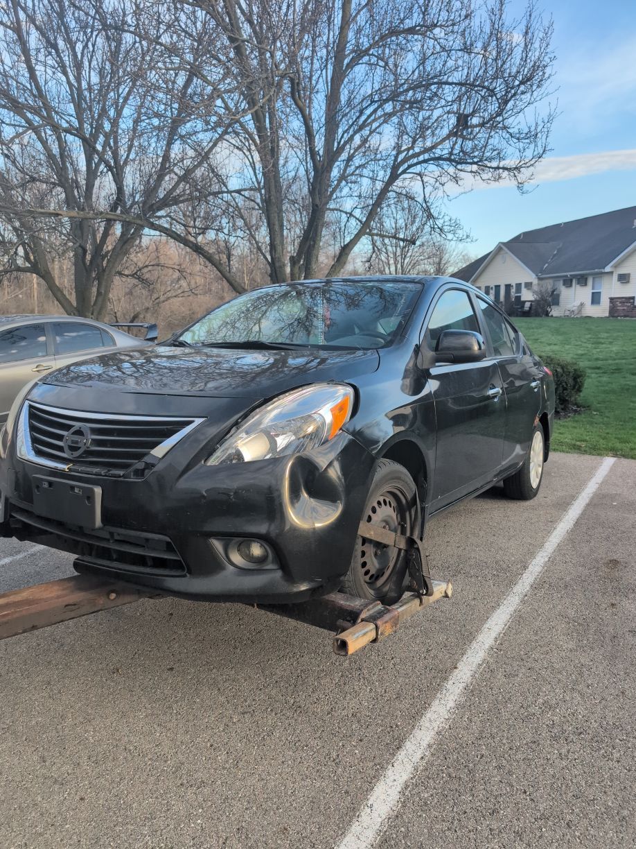 Damaged black sedan with missing front wheel parked in a lot with a wheel chock underneath