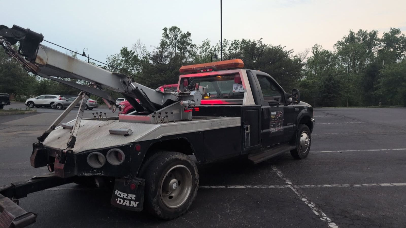Black tow truck with red lights parked in an empty lot, carrying a damaged car on its flatbed.