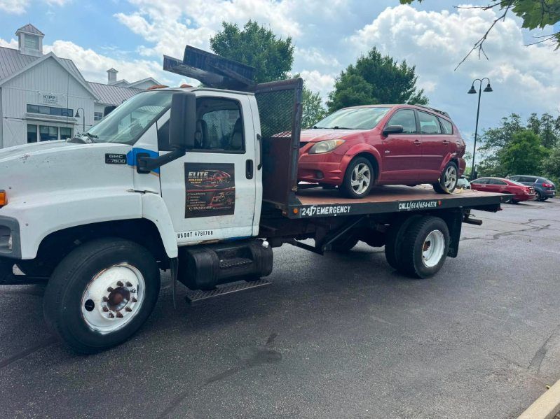 White flatbed tow truck carrying a red hatchback in a parking lot under a cloudy sky