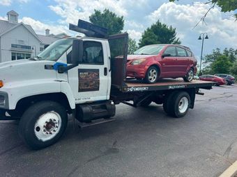 White flatbed tow truck carrying a red hatchback in a parking lot under a cloudy sky