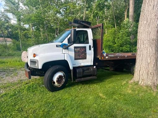 White flatbed truck parked on grass beside a large tree in a wooded area