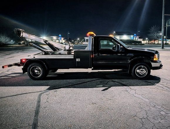 Black tow truck with flatbed and front-mounted wrecker parked in an industrial lot