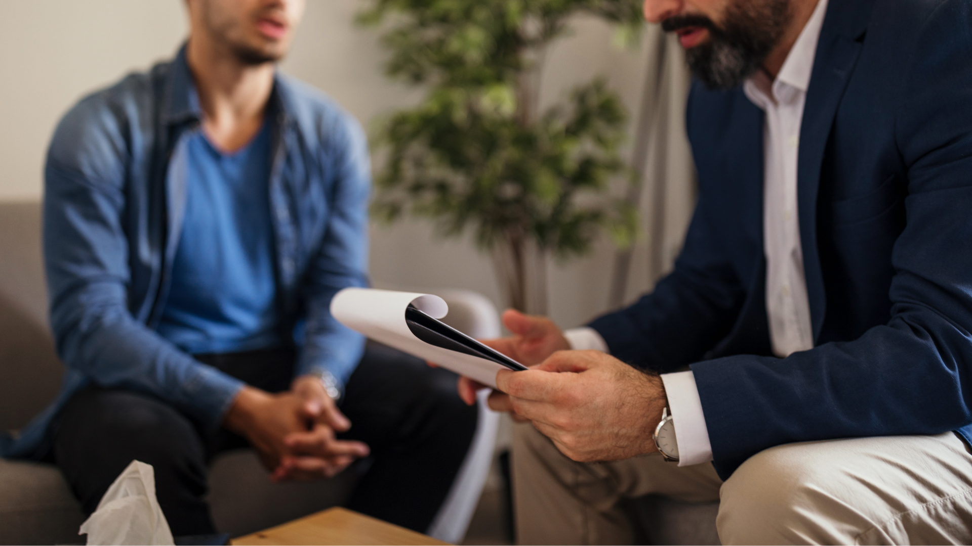 Two people in a meeting, one holding papers and talking while seated on a couch.