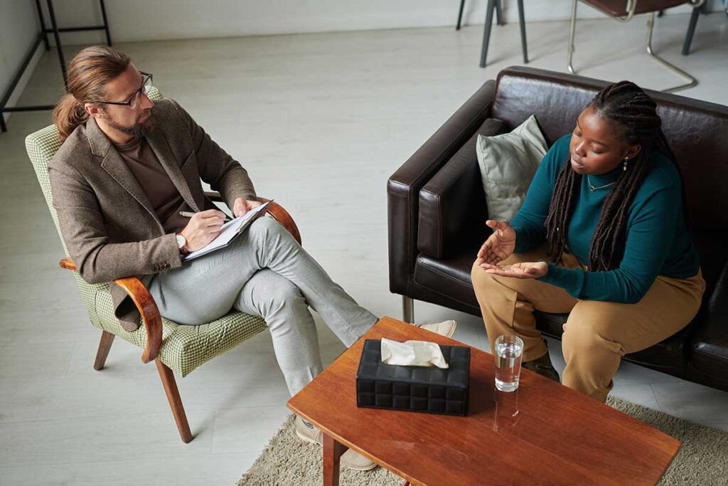 Two people in a counseling session, one taking notes while the other sits on a sofa speaking.