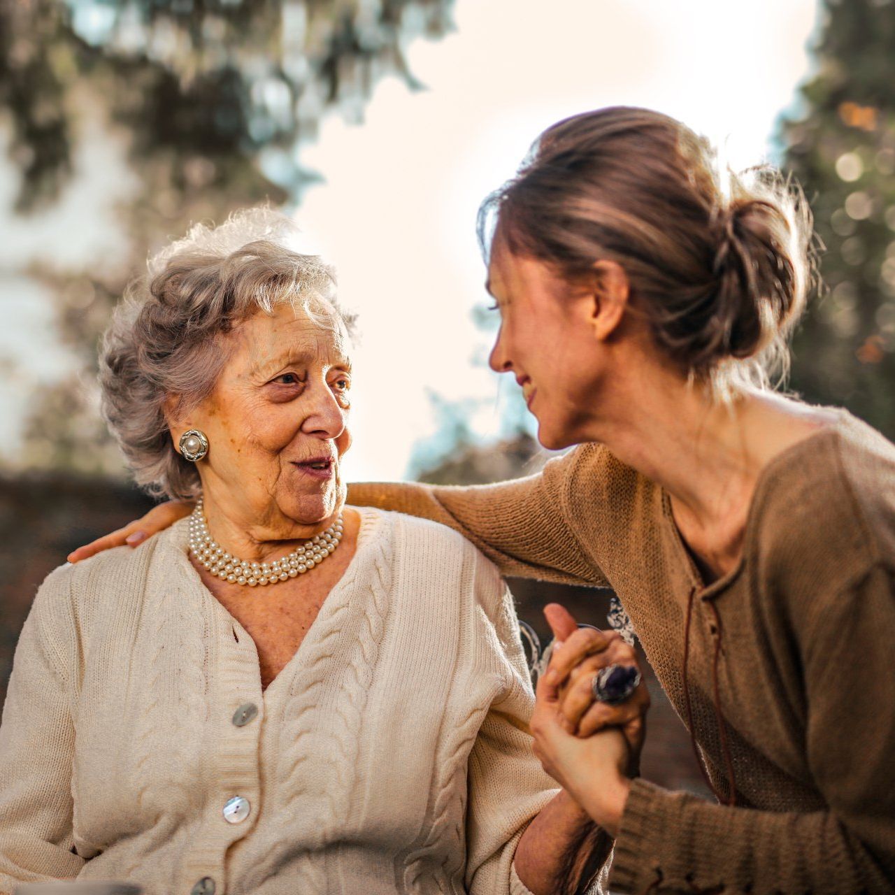 Woman comforting older woman outdoors; woman holds older woman's hand, touching her shoulder.