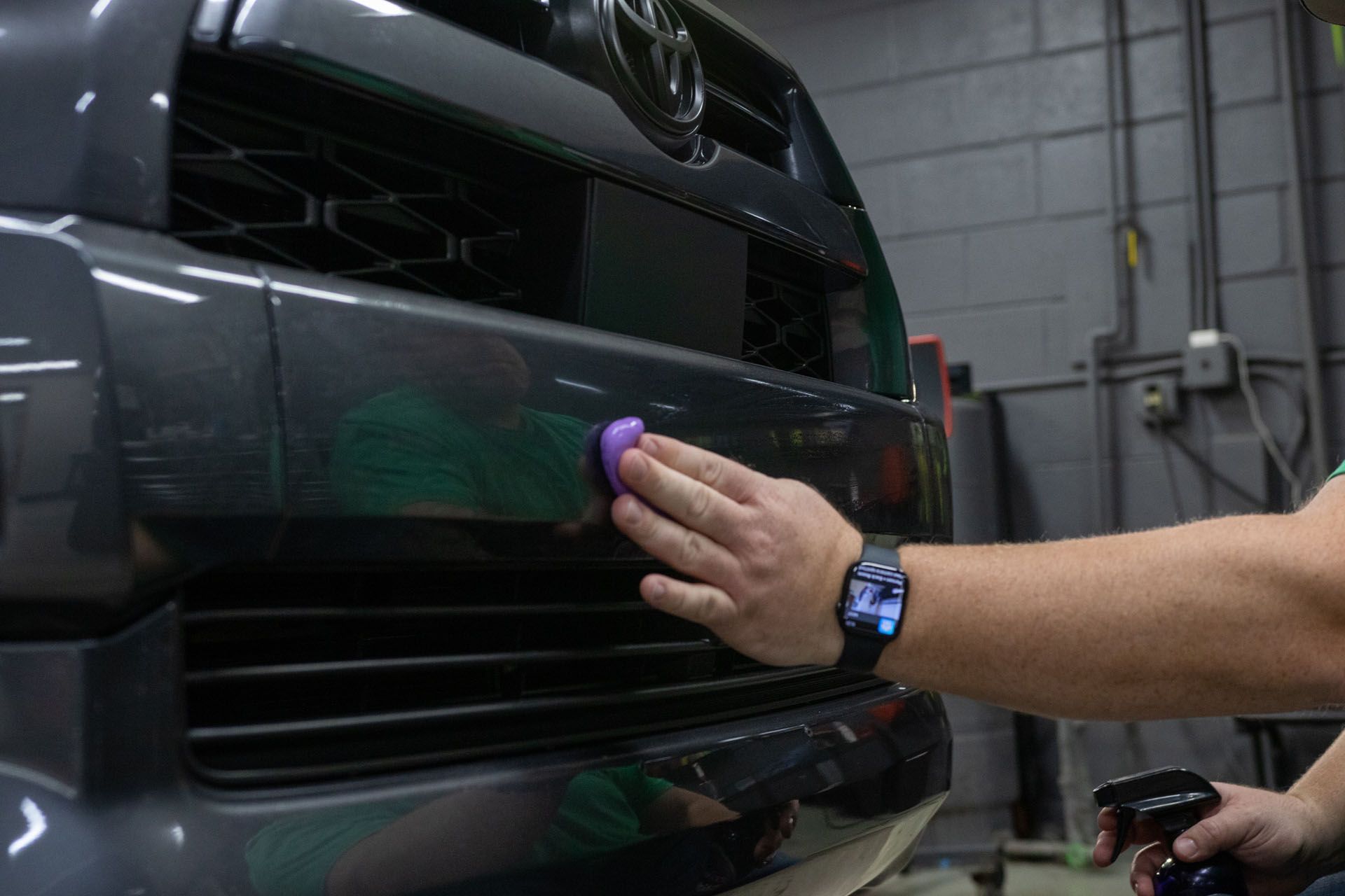 Hand wiping a glossy black car grille with a purple sponge in a garage