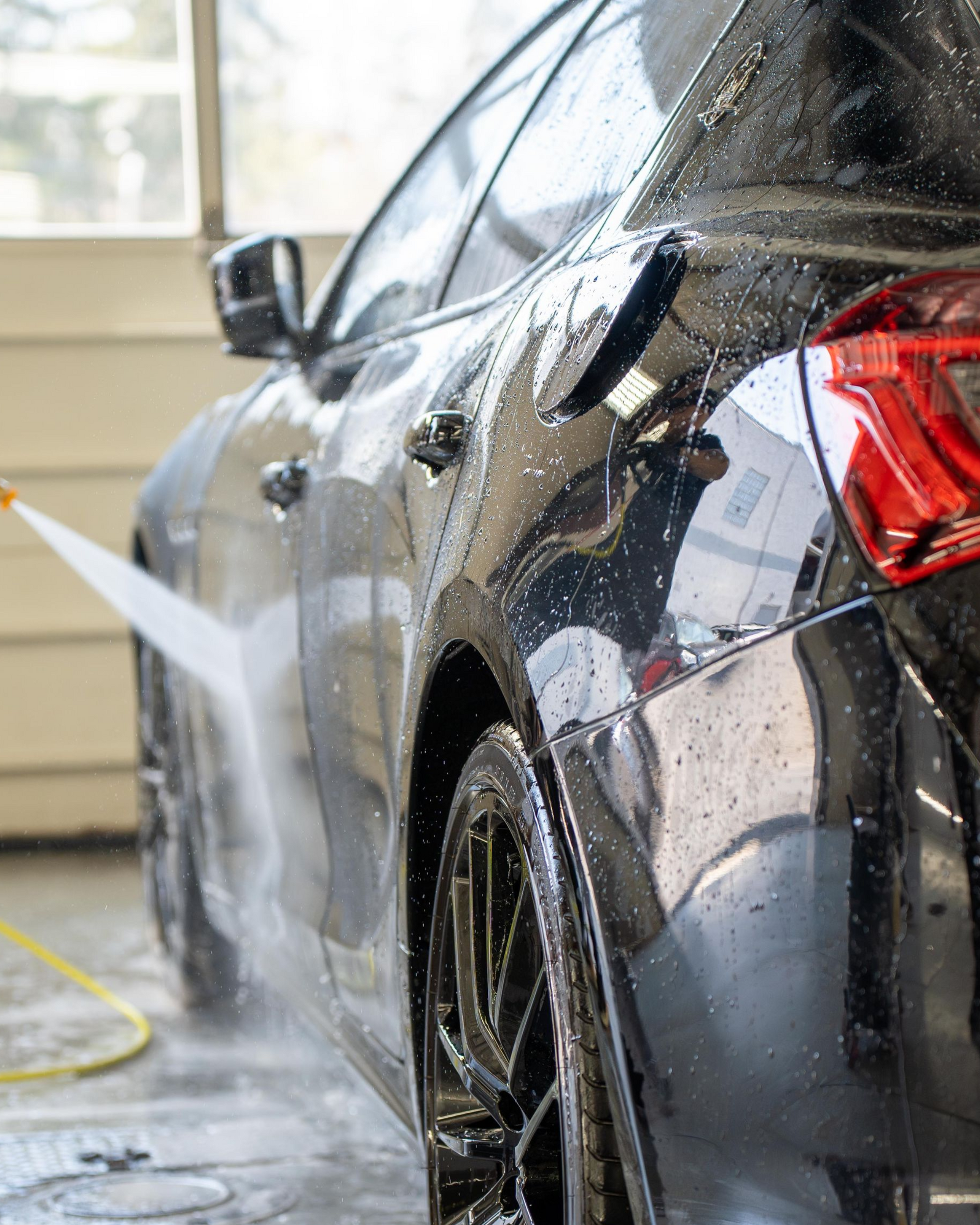 Black car being washed with a hose at a car wash, water spraying over the rear side.