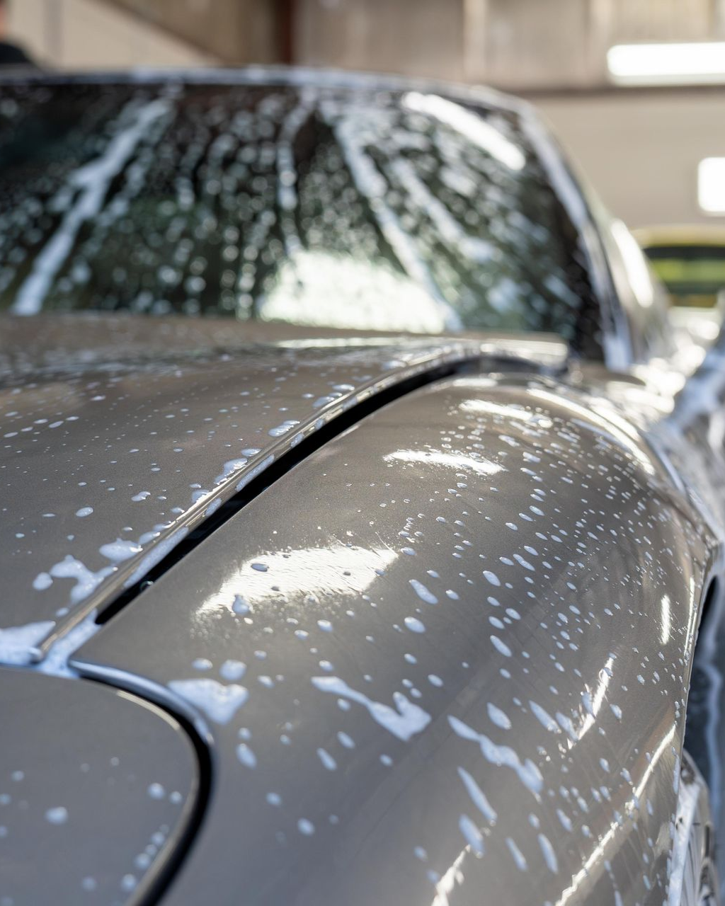 Close-up of a gray car hood and windshield covered in soap suds during a car wash