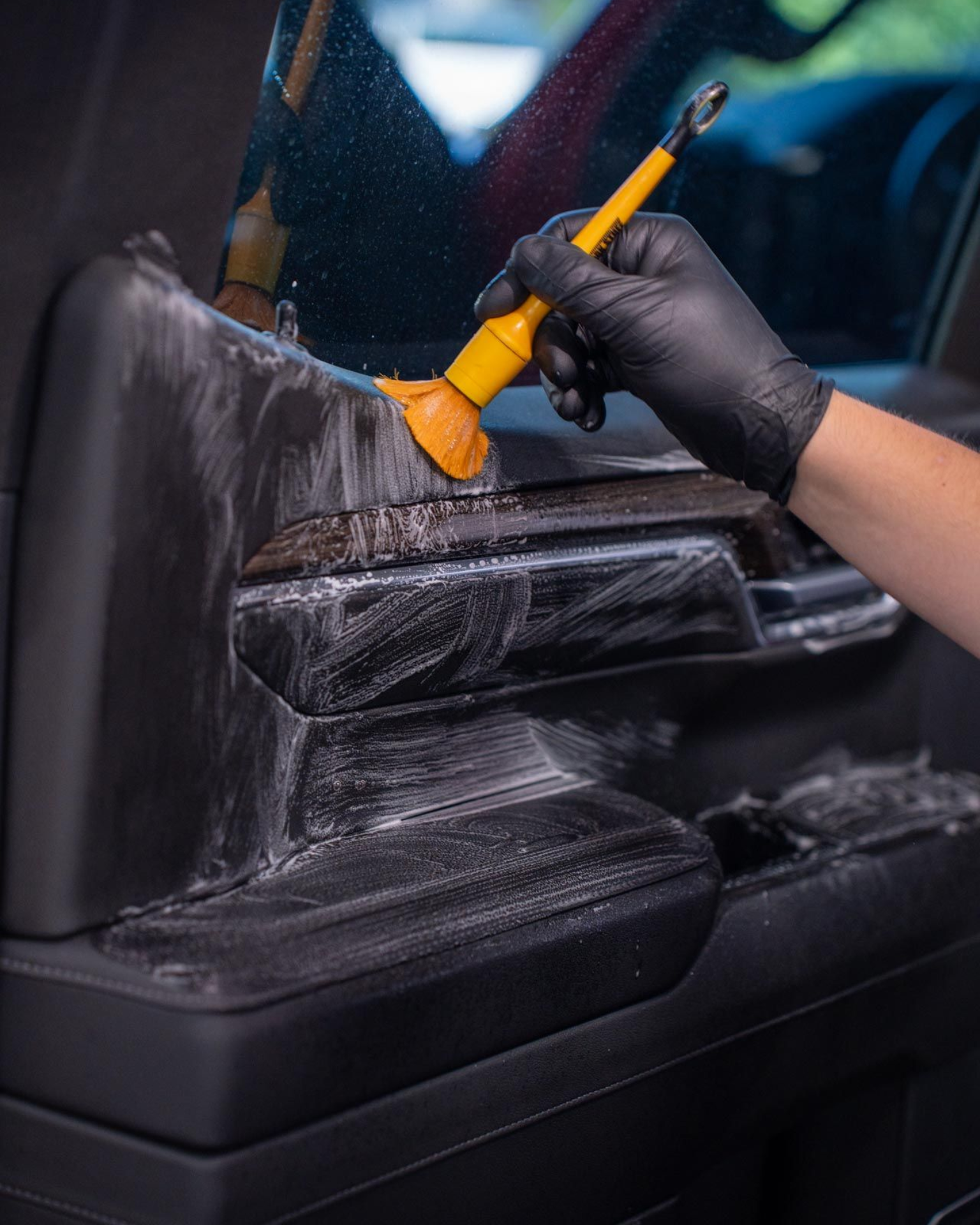 Gloved hand cleaning a black car seat with an orange brush and spray bottle.