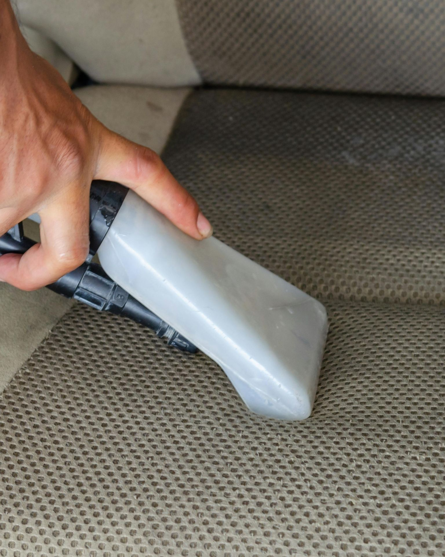 Hand holding a gray upholstery cleaning nozzle against a car seat.