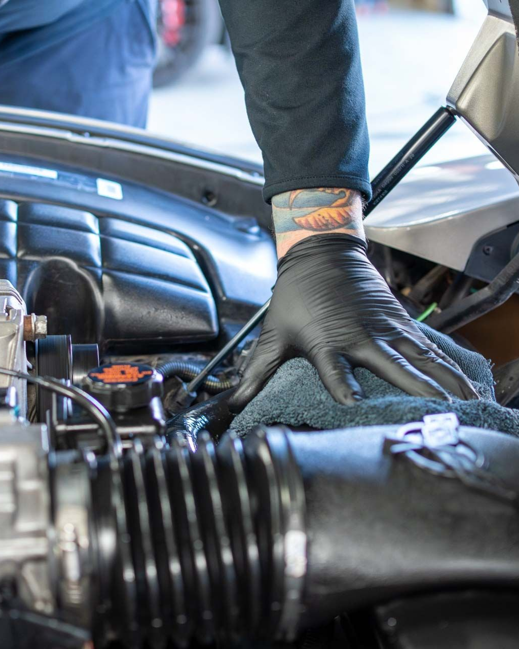 Black-gloved hand working inside a car engine bay beside a plastic air intake hose.