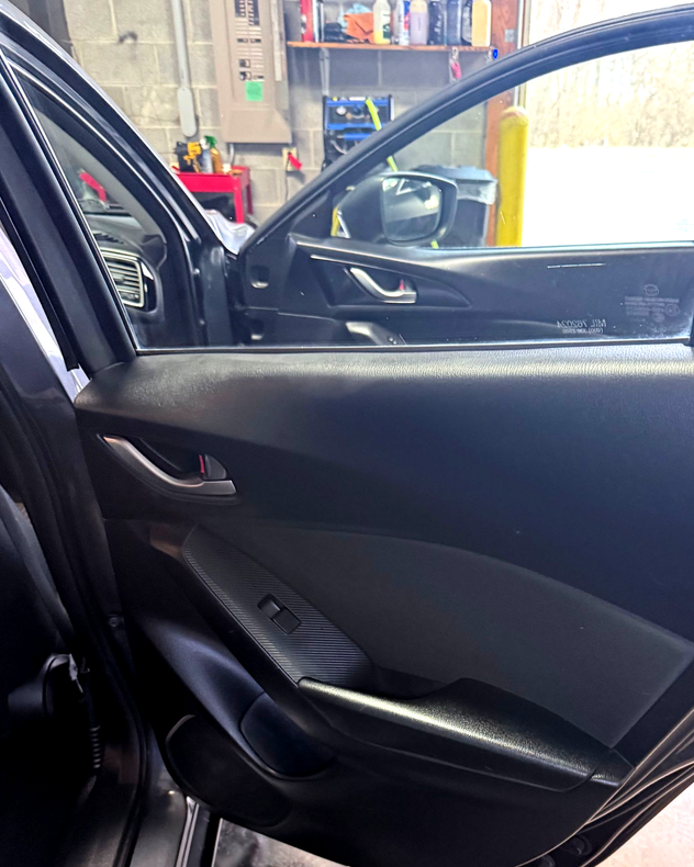 Car interior door panel in black leather, seen from the front seat with a workshop visible outside the window