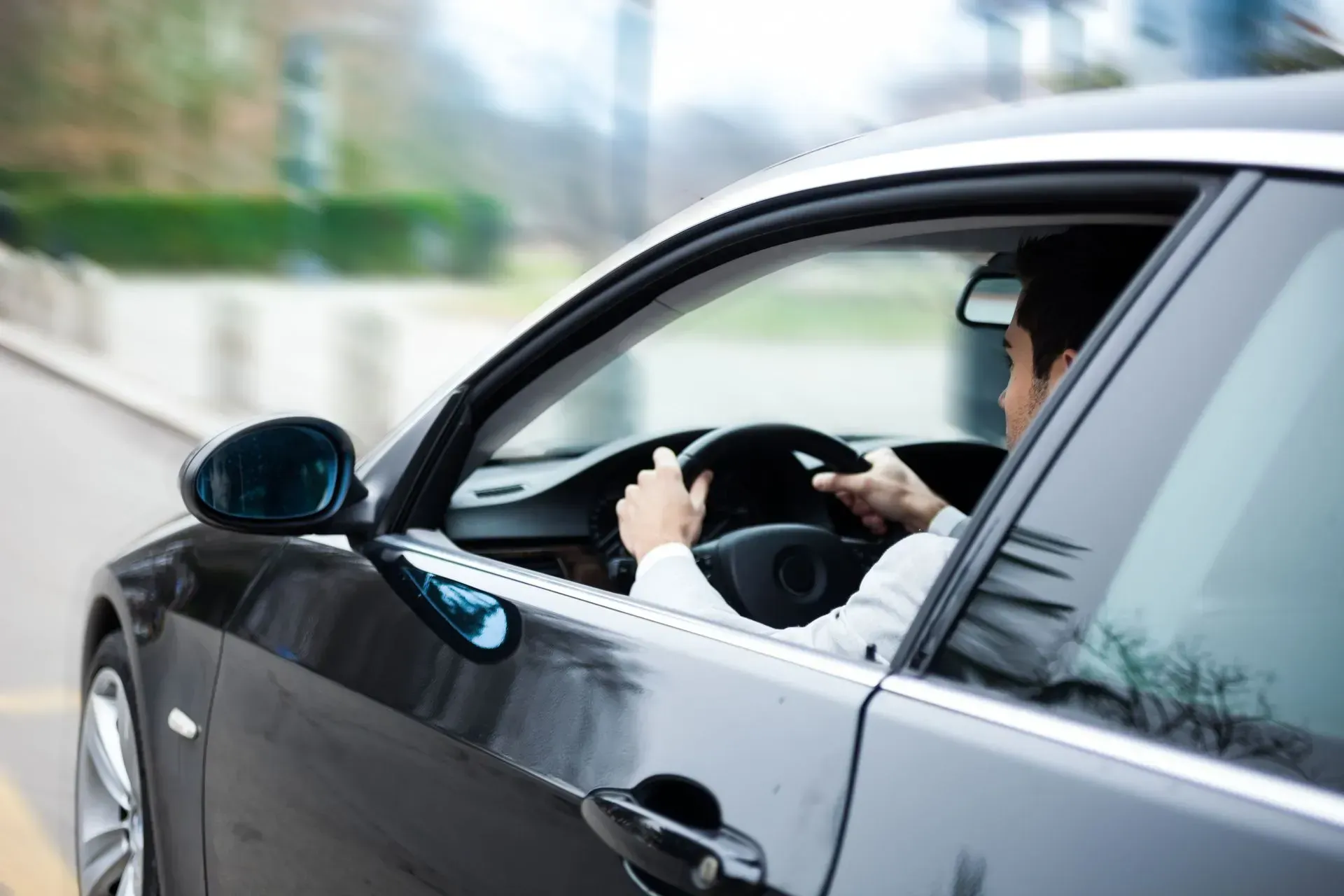 Person driving a gray car, seen through the open window on a blurred city street.