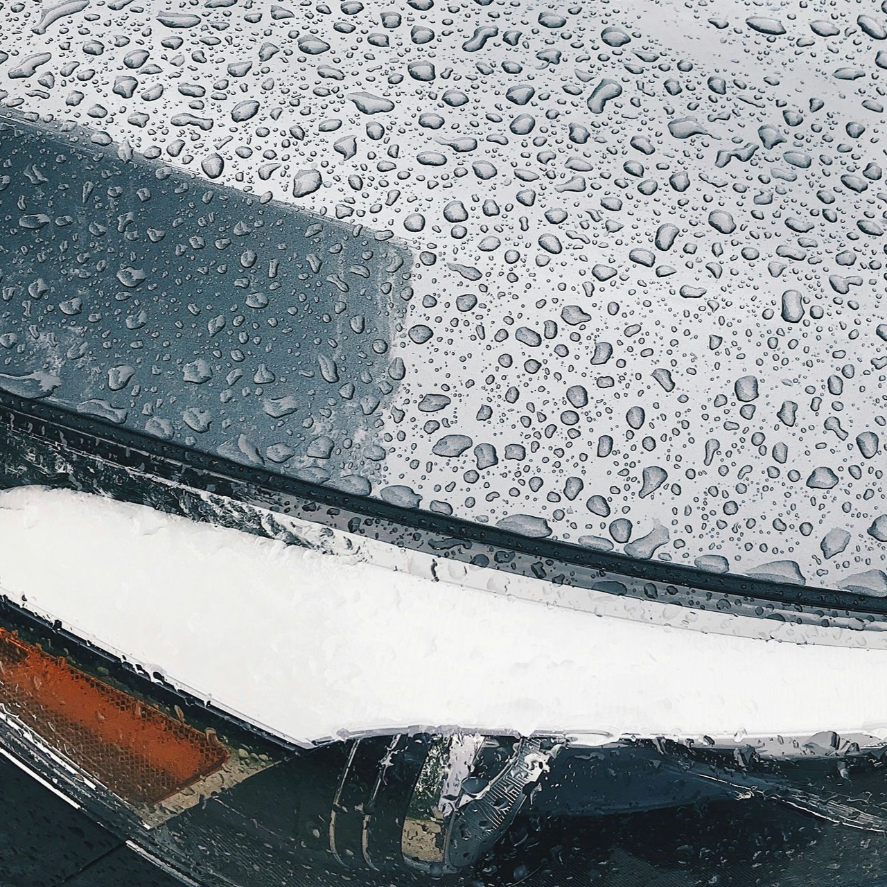 Close-up of a car hood covered in raindrops with a headlight and orange side marker visible